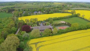 Paisaje rural con campos de flores amarillas, una casa con techo inclinado y una cancha de tenis.