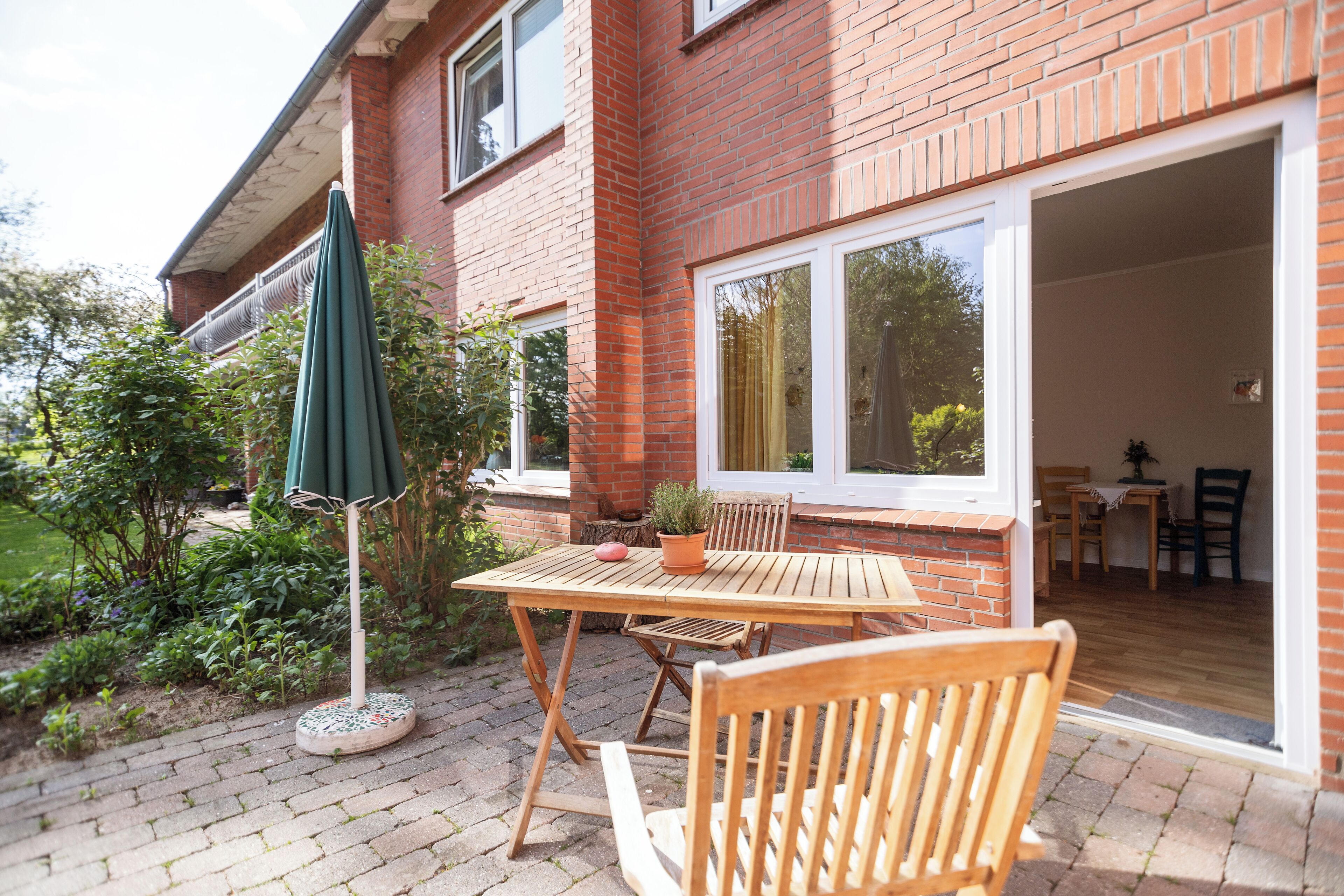 A patio with a wooden table and chairs, a green umbrella, and a brick house with a sliding glass door.