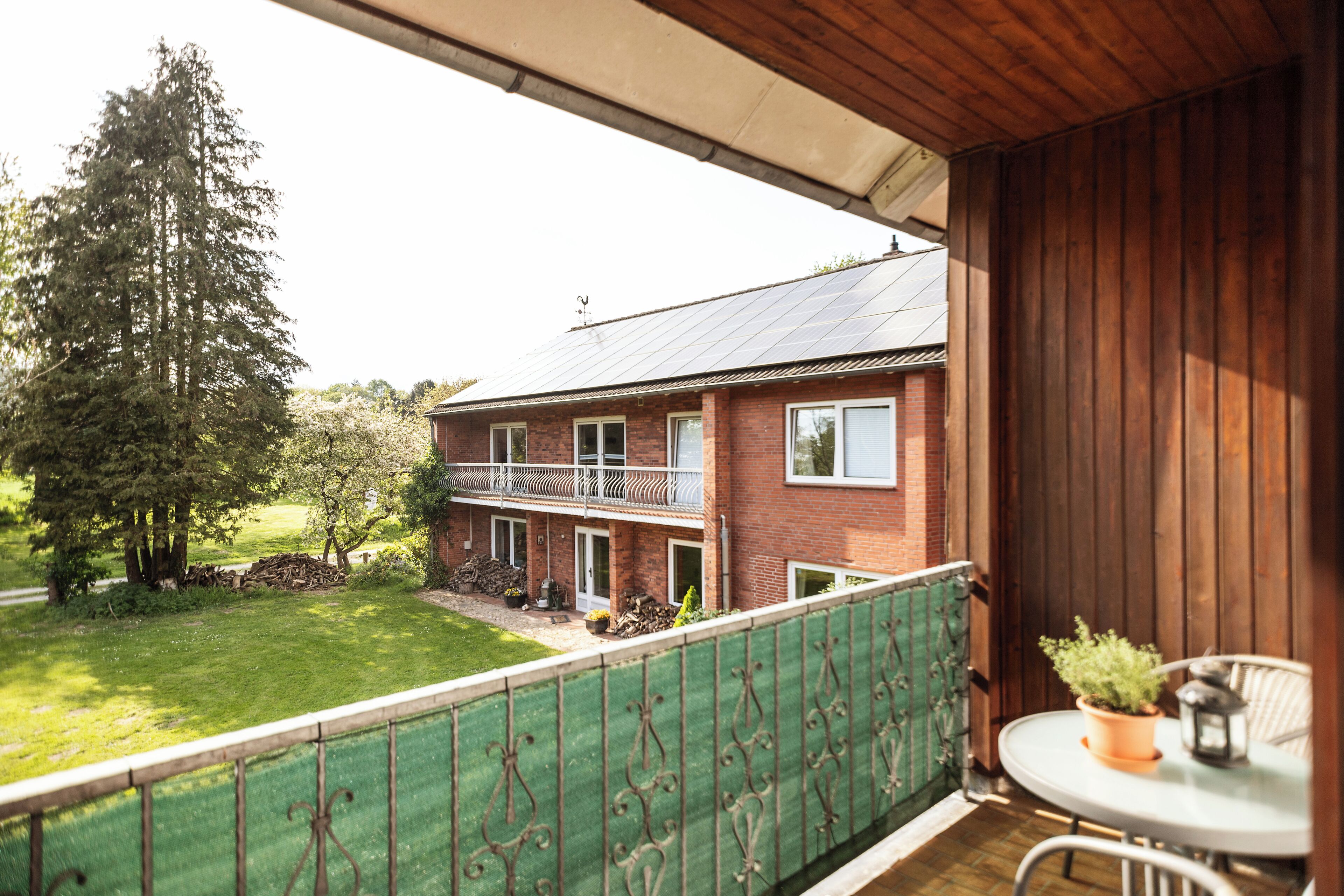 Ein Balkon mit grüner Metallreling, einem weißen Tisch mit einem Topfpflanzen und Blick auf ein Backsteinhaus mit Solardach.