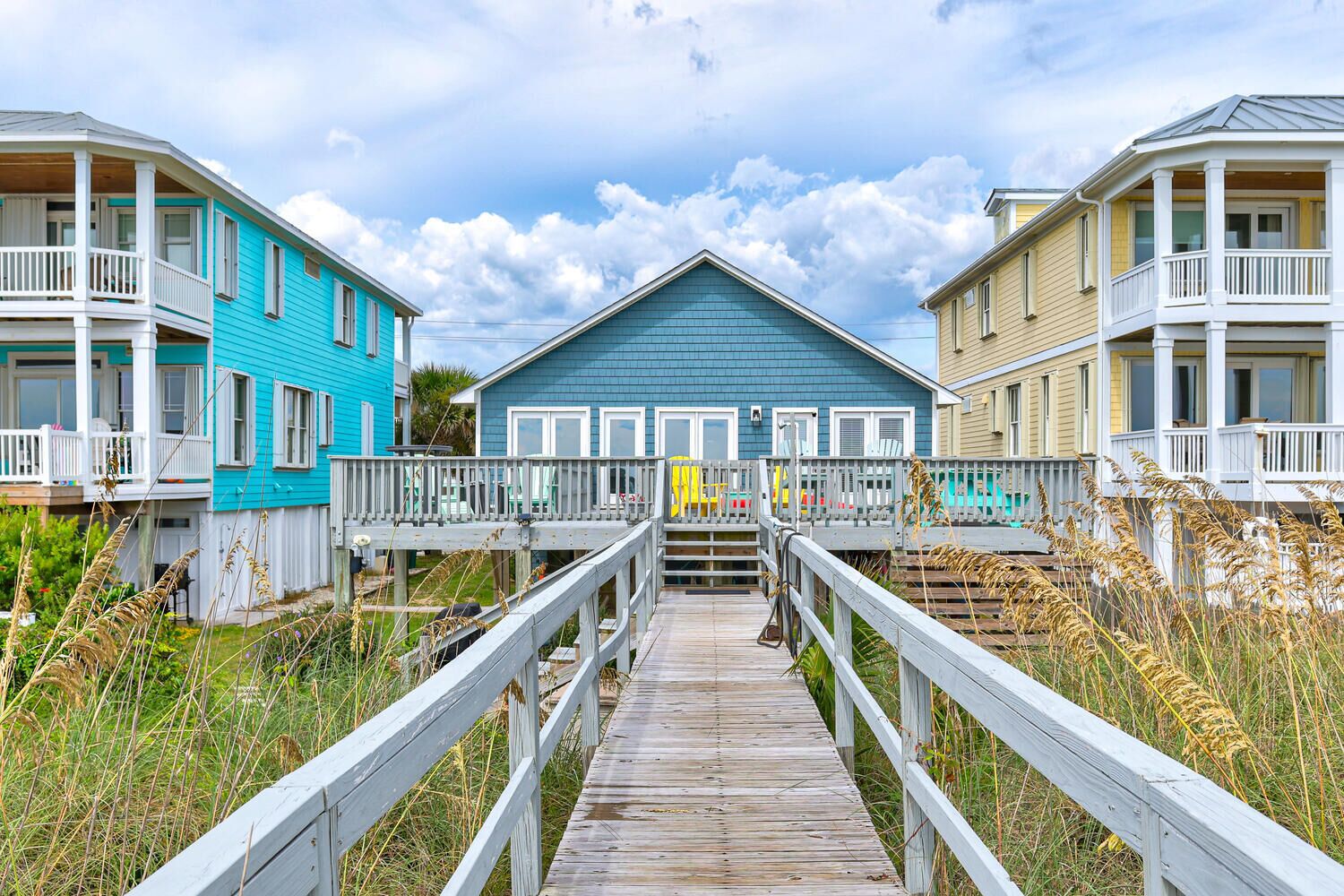 A wooden boardwalk leading to a row of beach houses with balconies and a blue house in the center.