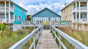 A wooden boardwalk leading to a row of beach houses with balconies and a blue house in the center. - Granny's Place - Oceanfront Home with Private beach access & Large Decks (Carolina Beach)