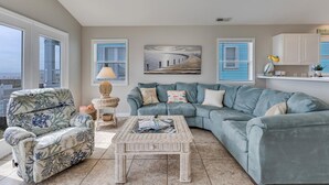 A living room with a large window, a blue sofa, a wicker coffee table, and a lamp. - Granny's Place - Oceanfront Home with Private beach access & Large Decks (Carolina Beach)