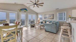 A spacious living area with a high ceiling fan, a dining table with chairs, a blue sofa, and a view of the ocean through large windows. - Granny's Place - Oceanfront Home with Private beach access & Large Decks (Carolina Beach)