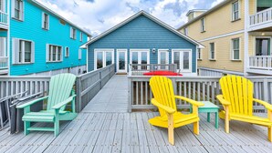 A deck with wooden railings and colorful Adirondack chairs, leading to a building with a blue facade and white doors. - Granny's Place - Oceanfront Home with Private beach access & Large Decks (Carolina Beach)