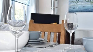 A dining table set with plates, bowls, and wine glasses, a wooden chair, and a painting of a coastal scene. - Spacious Modern Apartment with Sea Views in Prime Beachside Location (Westward Ho!)
