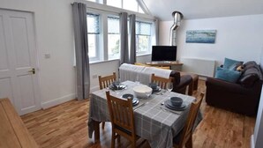 A dining area with a table set for four, a television on a stand, and a sofa in the background. - Spacious Modern Apartment with Sea Views in Prime Beachside Location (Westward Ho!)
