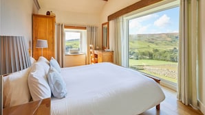 Een slaapkamer met een groot bed, een houten hoofdbord, een houten kledingkast, een nachtkastje, een lamp en uitzicht op glooiende heuvels door een groot raam.