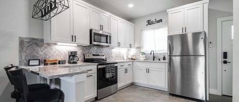 A modern kitchen with white cabinets, stainless steel appliances, a herringbone tile backsplash, and a bar area with stools.
