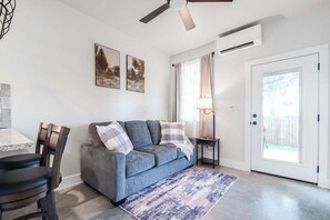 Ein Wohnzimmer mit grauem Sofa, einem Couchtisch und einem Essbereich mit Stühlen. Eine Deckenleuchte, eine Lampe und eine Tür zum Außenbereich sind vorhanden.