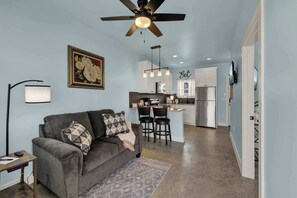 A modern living room with a gray sofa, a ceiling fan, and a kitchen in the background.
