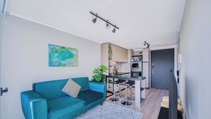 Ein modernes Wohnzimmer mit einem petrolfarbenen Sofa, einer kleinen Pflanze und einer Küchenzeile mit Barhocker.