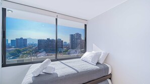 Ein Schlafzimmer mit großem Fenster und Blick auf die Stadt, einem Bett mit grauer Steudecke und weißen Kissen.