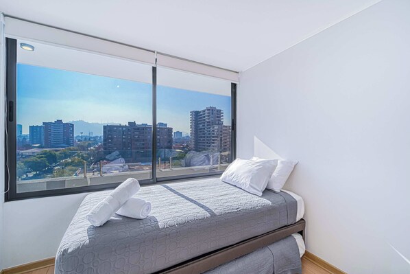 Un dormitorio con un ventanal que ofrece vistas de la ciudad, una cama con colcha gris y almohadas blancas.
