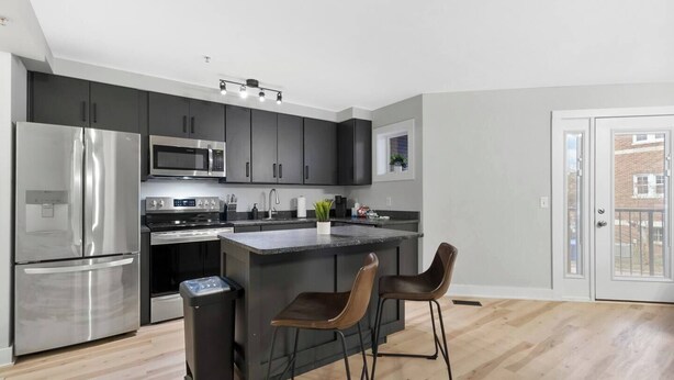 A modern kitchen with dark cabinets, stainless steel appliances, a central island with bar stools, and a view through a glass door. - The Lofts of Downtown 540 N 2nd St, #207 (Clarksville)