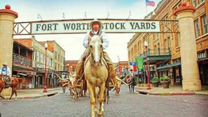 A street scene with a sign reading 'FORT WORTH STOCK YARDS' and a horse-drawn carriage in the foreground.