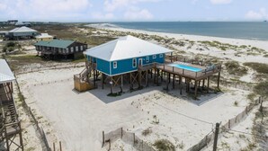 Una casa frente a la playa con piscina en una terraza de madera, rodeada de arena y vistas al océano.