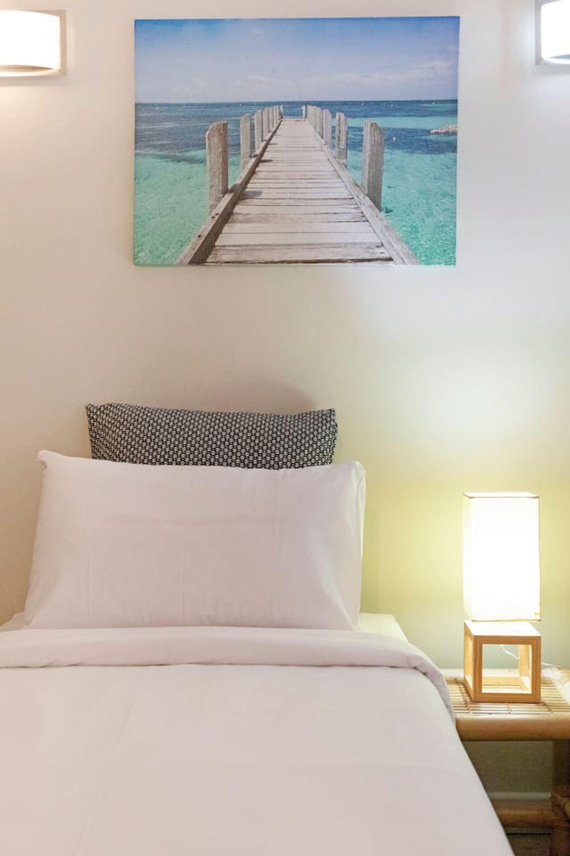 A bed with white bedding, a patterned pillow, and a wooden nightstand with a lamp. Above the bed is a framed picture of a wooden pier extending into turquoise waters.