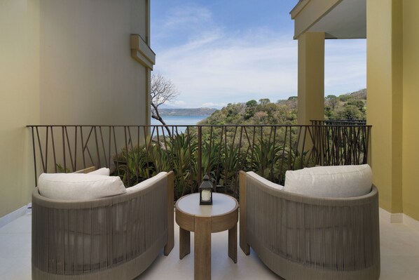 A balcony with two wicker chairs and a small table, overlooking a scenic view of a lake and hills.