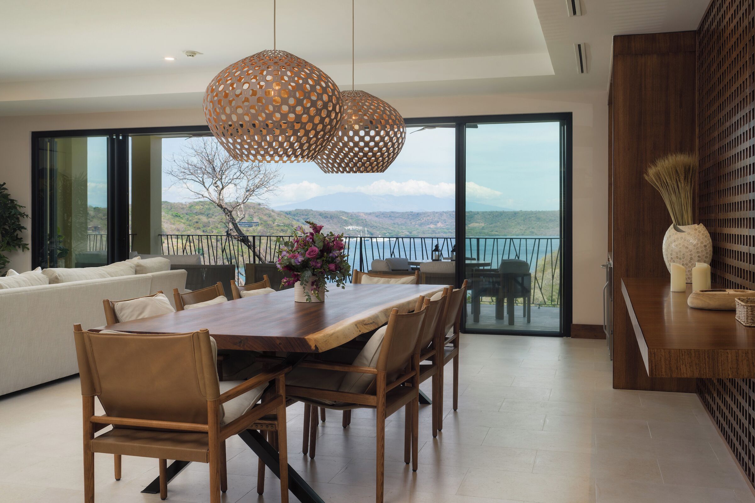 A modern dining area with a wooden table and chairs, a vase with flowers, and a view of the mountains through large glass doors.