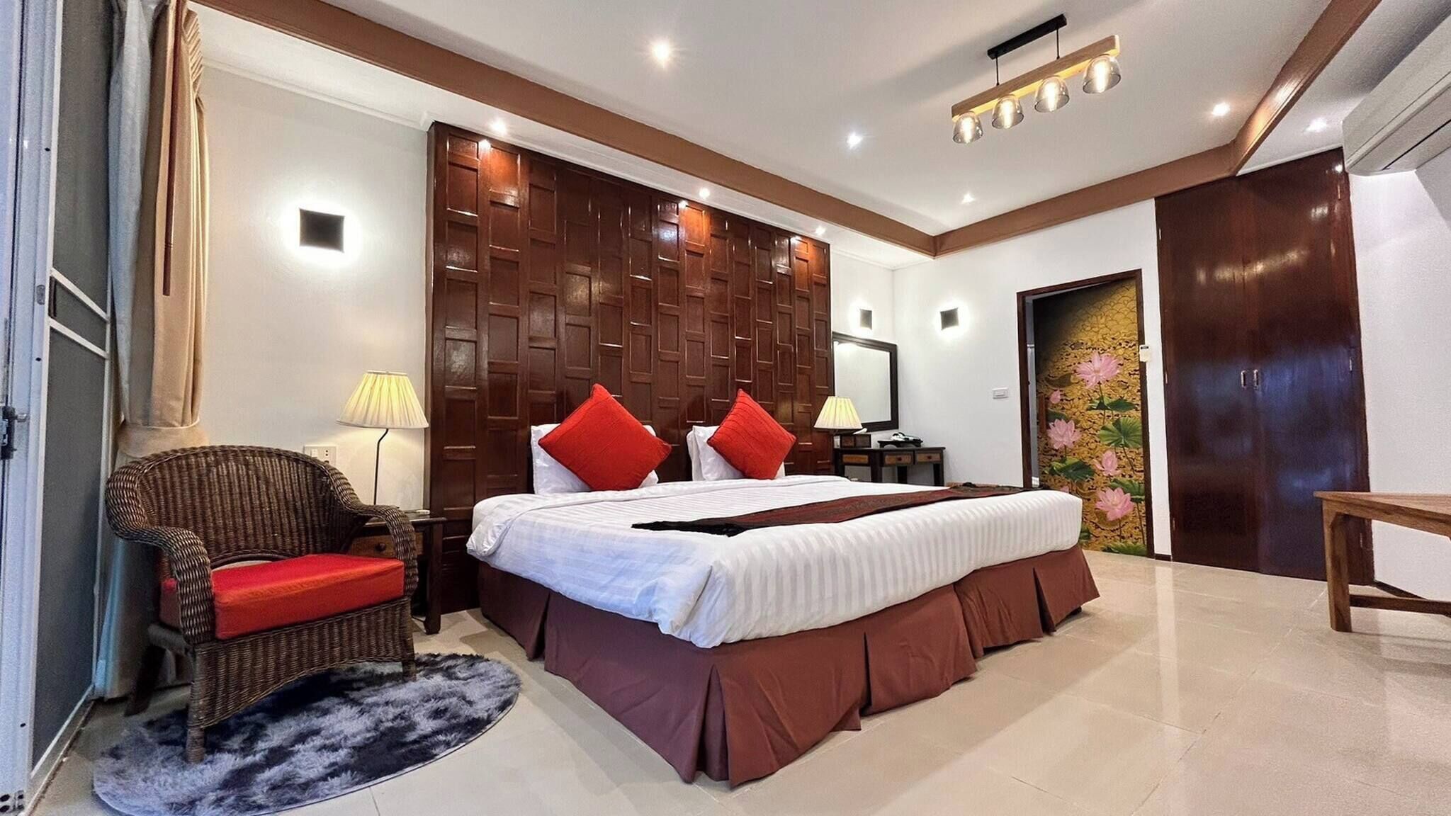 Una habitación de hotel con una cama grande, un cabecero de madera, una silla de mimbre, una mesita pequeña y una mesita de noche.