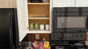 A kitchen with a black refrigerator, a microwave, and a stove. There are cabinets, a basket with snacks, and a shelf with kitchen items. - Trails, Tails, & Scales - This listing has two Cabins Rental (Burkeville)
