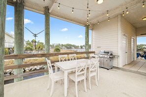 En täckt uteplats med ett vitt bord och stolar, en grill och utsikt över stranden och havet.