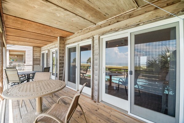 A covered outdoor area with a wooden deck, a round table with chairs, and large sliding glass doors offering a view of a pool and landscape.