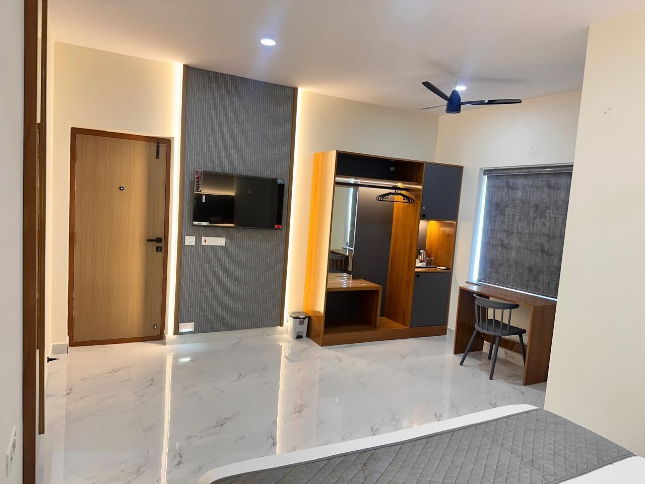 A modern hotel room with a wooden door, a flat-screen TV mounted on the wall, a small desk with a chair, and a large mirror.