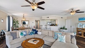 A spacious living room with a large sectional sofa, two ceiling fans, a dining area with a chandelier, and a kitchen in the background. - Updated Spacious condo with Coastal Decor across from beach with peeks of Gulf (Bradenton Beach)