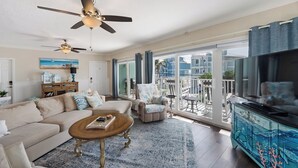 A spacious living room with a large sofa, a coffee table, and a TV stand. There are ceiling fans, a balcony with a view of the neighborhood, and a large window. - Updated Spacious condo with Coastal Decor across from beach with peeks of Gulf (Bradenton Beach)