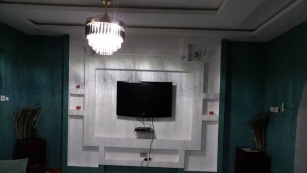 Una sala de estar con un televisor de pantalla plana grande montado en una pared blanca, una alfombra estampada y dos sofás de color oscuro con cojines.
