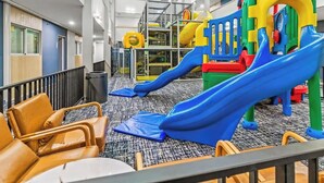 A children's play area with slides and climbing structures, a seating area with chairs, and a carpeted floor.