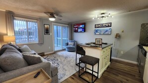A compact living space with a sofa, a small kitchen area, a dining table, and a TV mounted on the wall displaying a streaming service interface.
