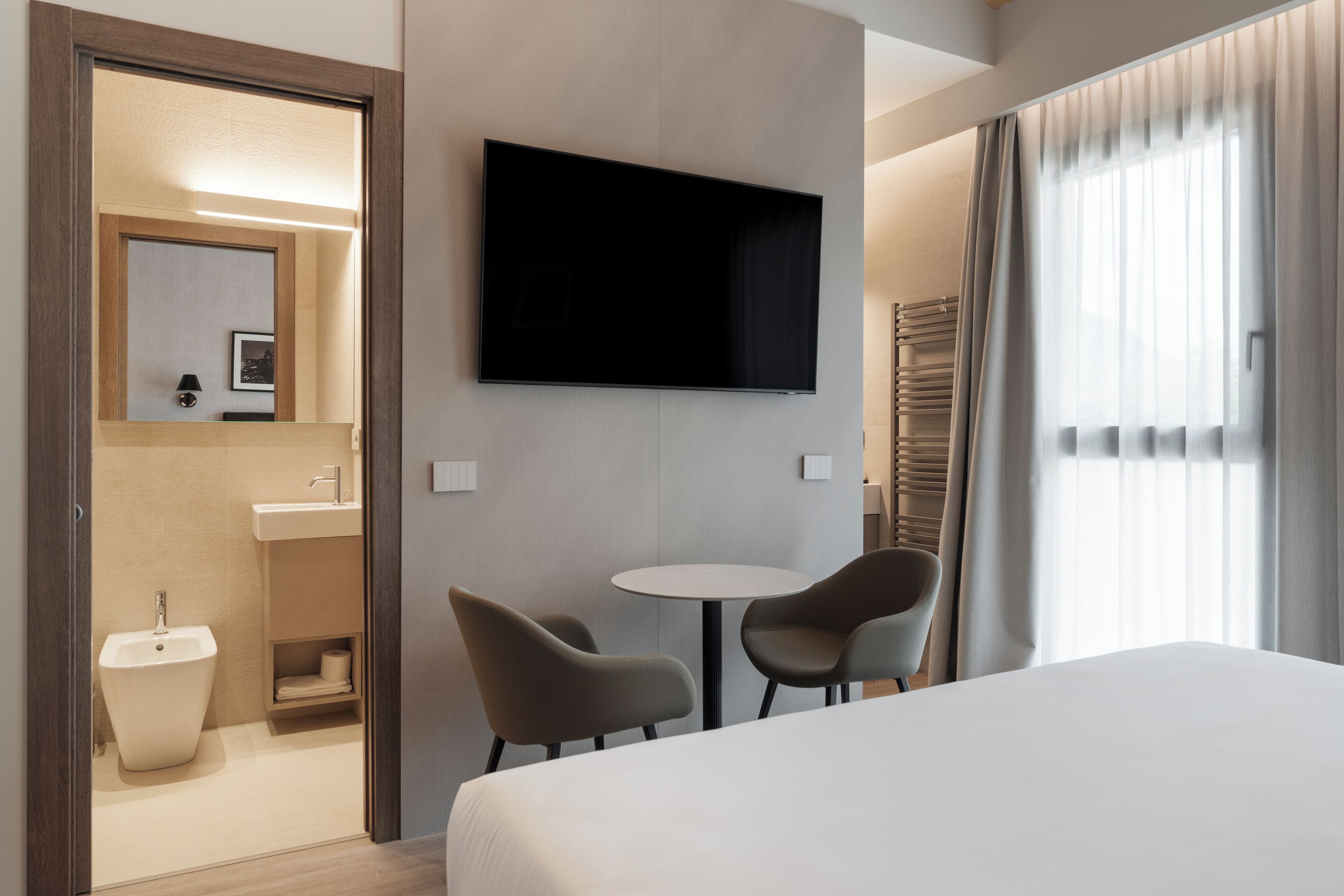 Habitación de hotel con cama, televisor montado en la pared, una mesita redonda con dos sillas, lavamanos y espejo.