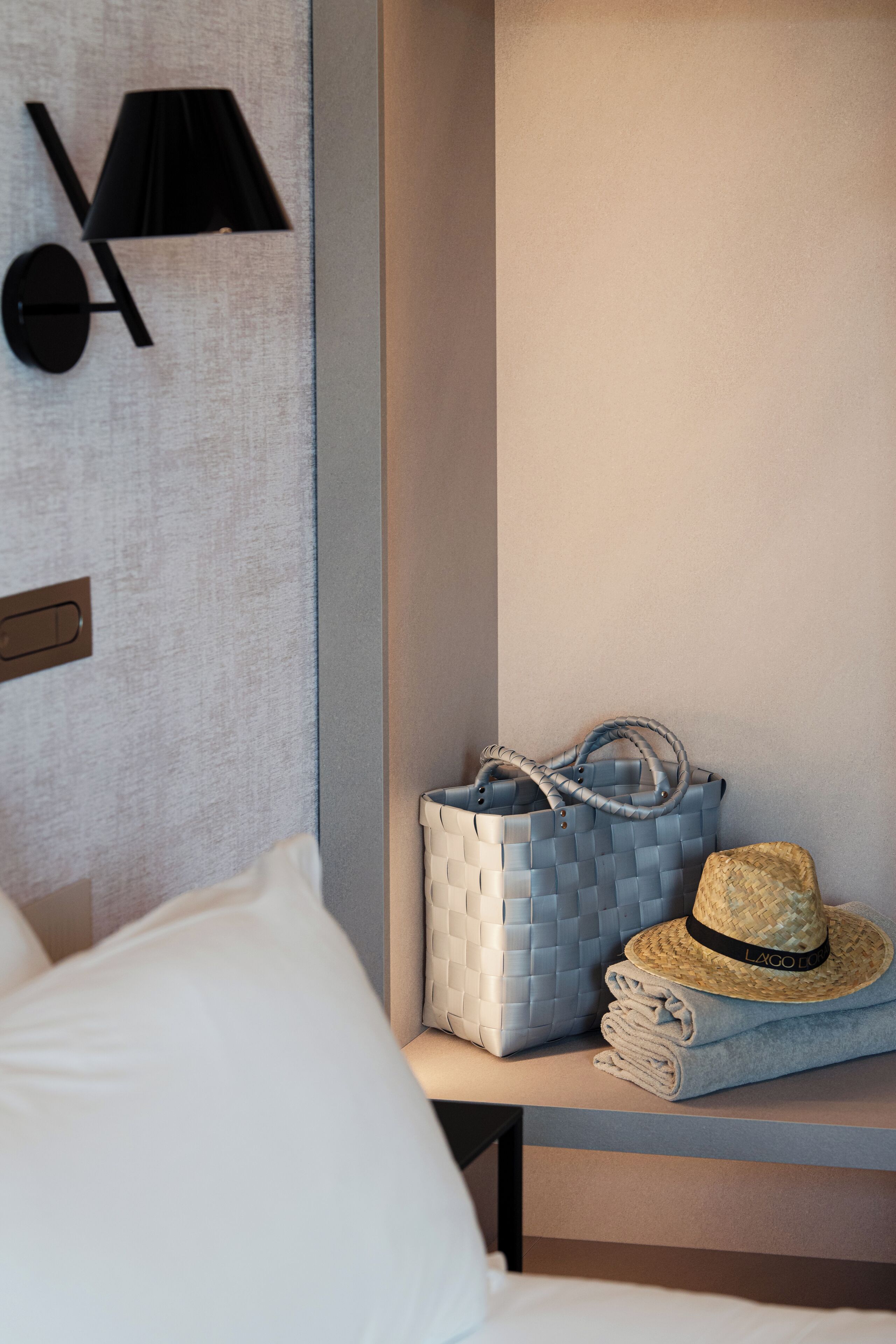 A neatly arranged hotel room with a bed, a wall-mounted lamp, a woven bag, and a straw hat on a small table.