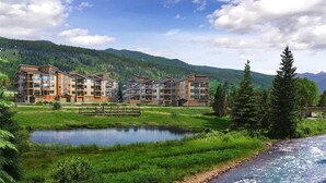 Un hôtel à plusieurs étages, doté d’une architecture moderne, situé au bord d’une rivière et offrant une vue sur une chaîne de montagnes boisée.