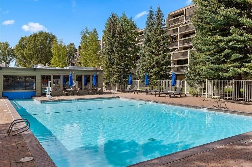 Una piscina con sillas de descanso y sombrillas, rodeada de árboles y un edificio con balcones.