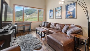 A modern living room with a large window, a brown leather sofa, a wooden coffee table, and a television. - Fp300 Foxpine Inn 2 Bedroom Condo (Copper Mountain)