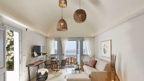 Un salon moderne avec un canapé, un pouf et un coin repas. On y trouve des suspensions en osier, un téléviseur à écran plat et une vue sur la mer grâce à de grandes fenêtres.