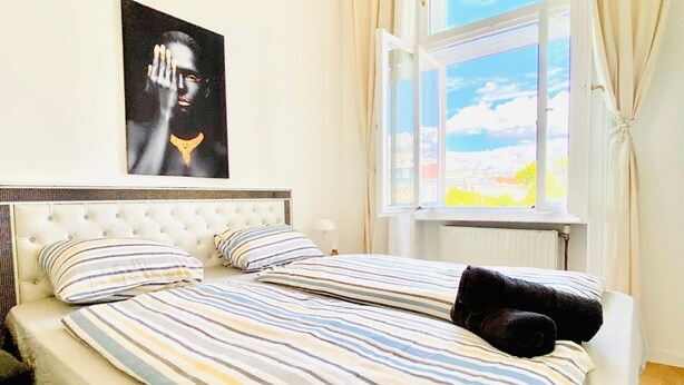 A double bed with striped bedding, a tufted headboard, a window with curtains, and a poster on the wall. - Cozy Stay in Shared Apt  Short Walk to Westbahnhof (Vienna)