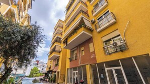Un edificio residencial amarillo de varios pisos con balcones, una entrada visible en la planta baja y un árbol en primer plano.