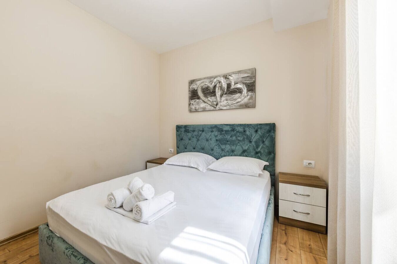 A neatly made bed with white linens and rolled towels, a teal upholstered headboard, and a single artwork above it.