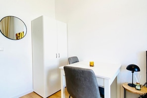 Quarto com uma mesa branca, uma cadeira cinzenta, um guarda-roupa branco, um espelho redondo e uma pequena mesa com abajur e garrafa.