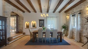 A dining area with a stone wall, wooden beams, a chandelier, and a dining table with chairs.