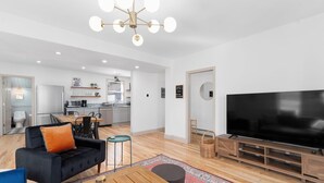 Una sala de estar moderna con un sofá negro, una mesa de centro de madera, un televisor plano grande y una cocina al fondo.