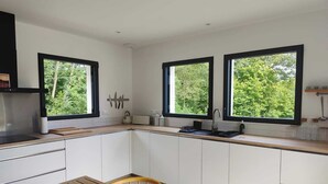 A modern kitchen with white cabinets, a wooden table, and large windows offering a view of greenery. - Villa in the southern moors, 10 minutes from the ocean (Magescq)