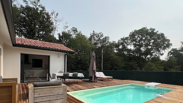 A wooden deck with a swimming pool, a small building with a tiled roof, and outdoor furniture. - Villa in the southern moors, 10 minutes from the ocean (Magescq)