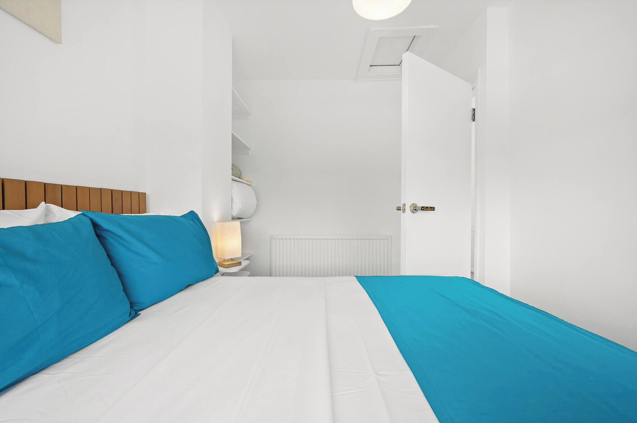 A bedroom with a bed, white walls, a wooden headboard, a nightstand with a lamp, and a door leading to another room.