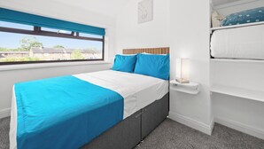 A bedroom with a bed, a bedside table with a lamp, a window with a view of buildings, and a shelf with a pillow.