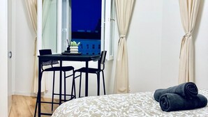 A room with a bed, a table with two chairs, a window with curtains, and a view of buildings outside.